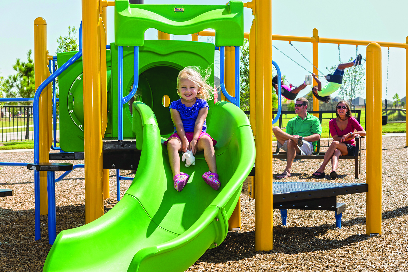 A child in a blue shirt and pink shoes slides down a green slide at a playground within the master-planned community, while two adults sit in the background and other children use swings. This vibrant area offers new homes near Rosenberg, perfect for growing families.