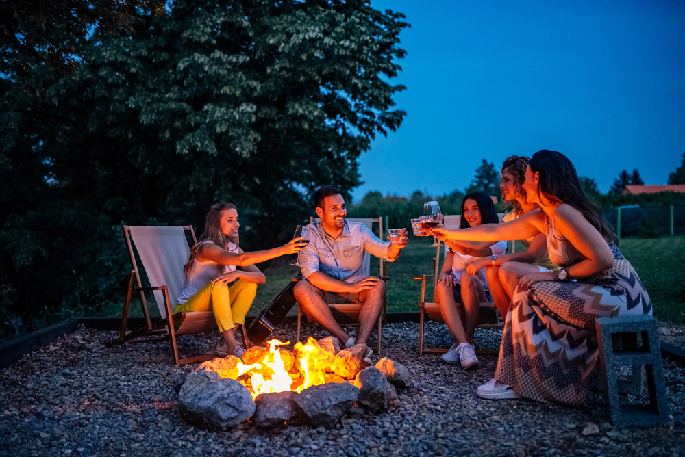 A group of people sitting around a campfire at dusk, toasting with drinks. They are outdoors on a gravel surface with trees in the background, enjoying the serene atmosphere of a master planned community near Richmond.