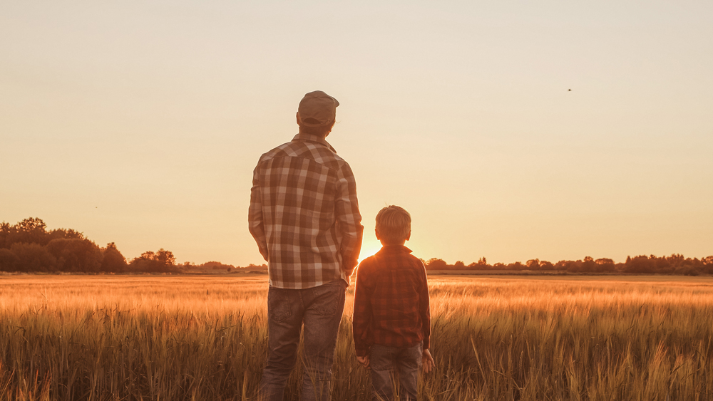 Two individuals, one adult and one child, wearing plaid shirts, stand side by side in a field at sunset near Rosenberg in Fort Bend County, gazing at the horizon.