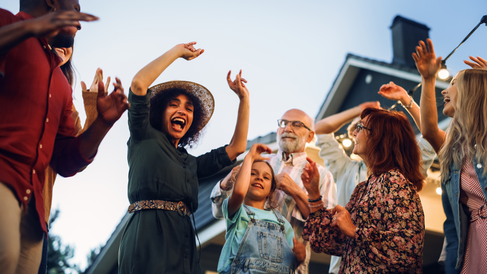 A group of people of various ages, including a child, are gathered outdoors near Richmond, smiling, dancing, and raising their hands in celebration. A house with lights is in the background.