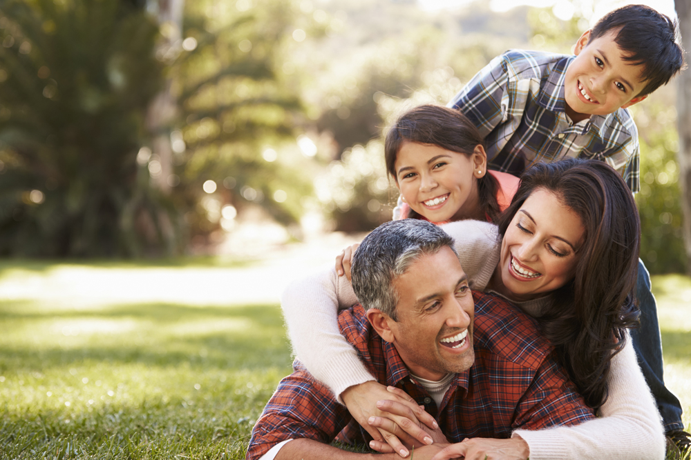 A joyful family of four, comprising two adults and two children, smiling and lying on top of each other on a grass lawn in a park within a master planned community in Fort Bend County.