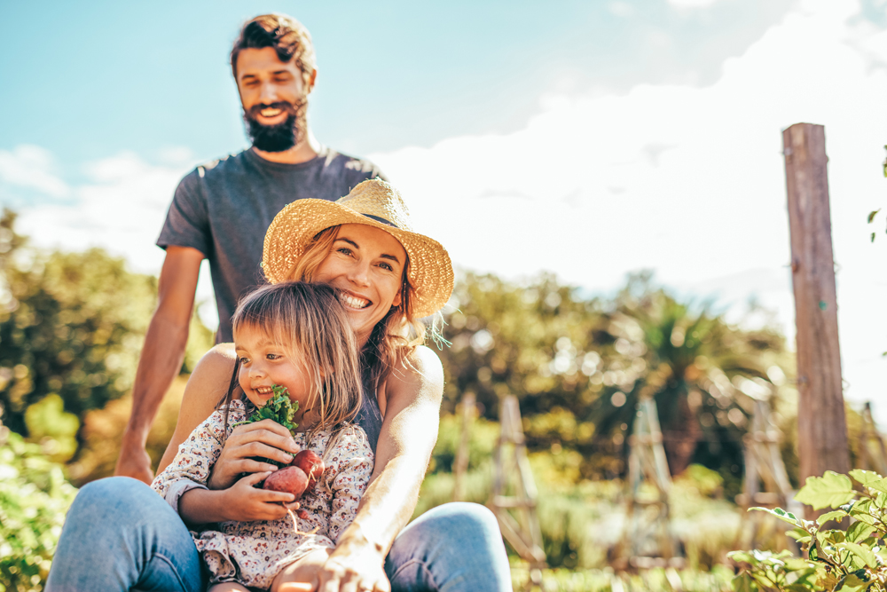 A smiling woman in a straw hat holds a child with short hair and vegetables, while a bearded man stands behind them in a garden under a bright sky near Richmond.