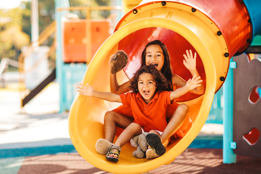 Two children, Emberly and a boy, slide down a yellow playground tube slide near Richmond, both smiling and extending their arms outward. The boy holds a stuffed bear. Bright playground equipment is in the background.