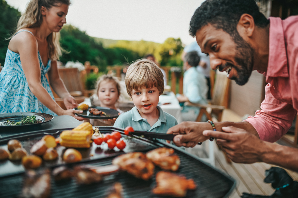 A man is grilling food as a young boy watches, near Richmond in Fort Bend County. A woman and another child are in the background. The grill is loaded with various foods, including corn, potatoes, and skewers. They are outdoors by one of the area