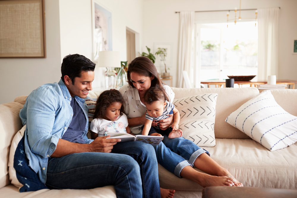 A family of four sits on a couch in a living room. The father reads a book to the two children while the mother looks on, all enjoying their new home near Rosenberg in Fort Bend County. The room is bright with natural light coming through the windows.