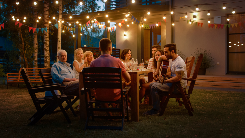 In a master planned community near Rosenberg in Fort Bend County, a group of people is seated around a wooden table in a backyard at night, with string lights overhead. One person is playing a guitar.