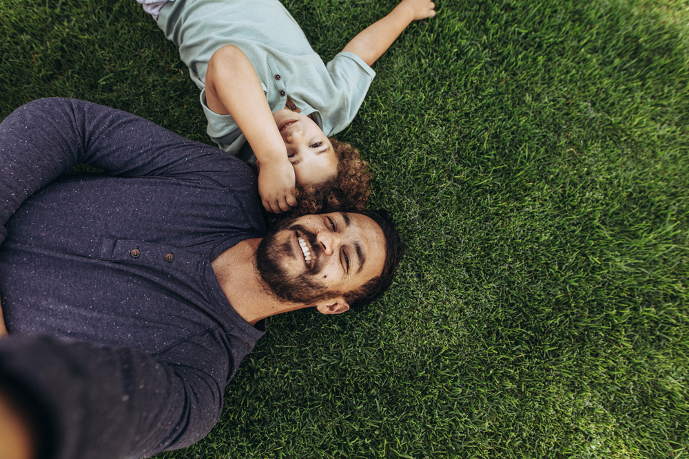 A man and a child lying on grass near Richmond, smiling up at the camera while capturing a selfie in Fort Bend County