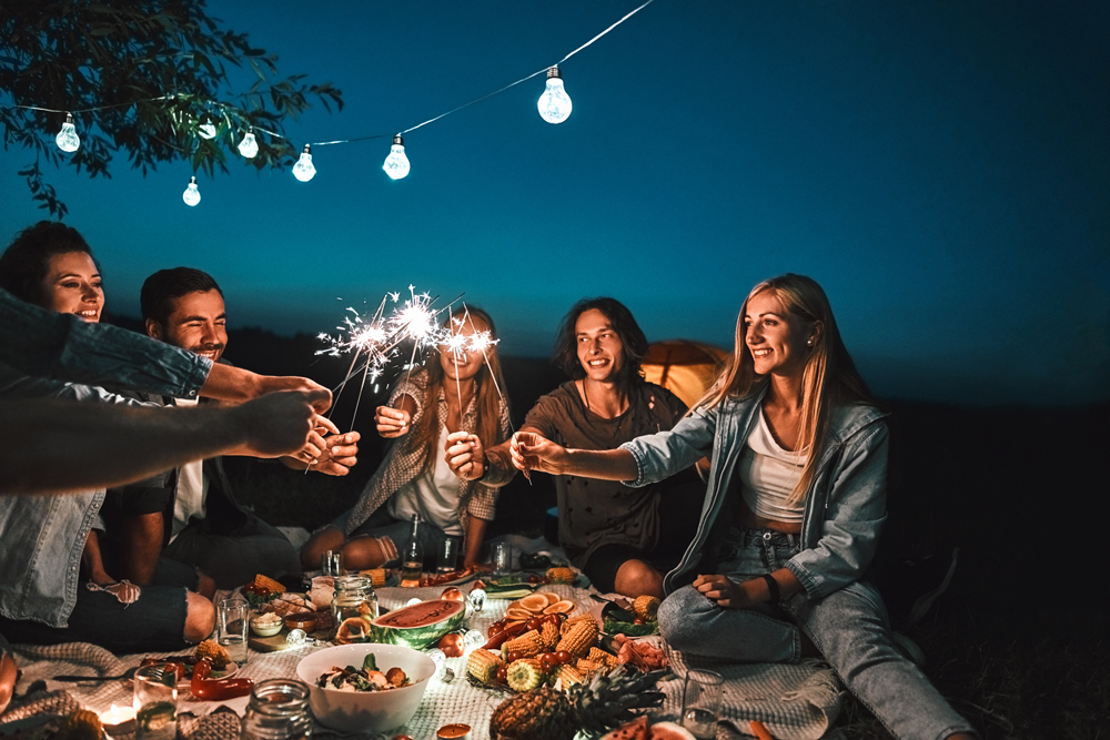 A group of people enjoying an outdoor evening picnic near Richmond, holding sparklers under fairy lights strung above. Within this master planned community, the picnic spread includes various foods and drinks.