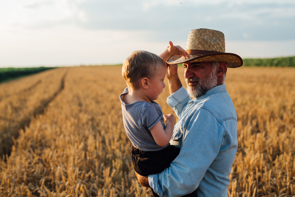 An adult holding a child stands in a wheat field at sunset, just outside the master planned community of Emberly near Richmond. The adult adjusts their straw hat while looking lovingly at the child.