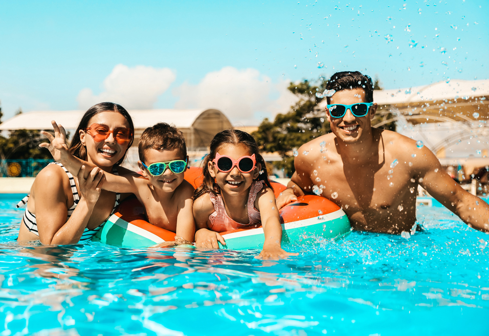 A family of four wearing sunglasses and swimsuits enjoys splashing in a swimming pool on a sunny day in the master planned community of Emberly near Rosenberg.