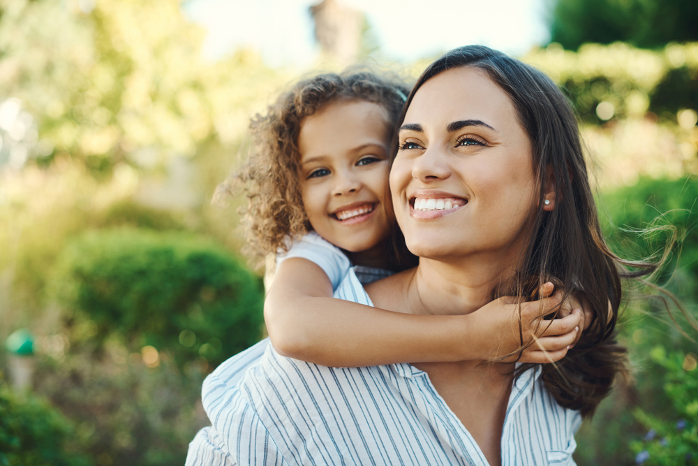 A woman is smiling while a child with curly hair hugs her from behind in an outdoor setting near Rosenberg, with greenery in the background.