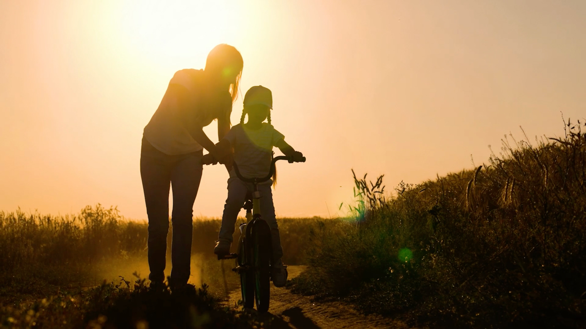 A woman helps a child ride a bike on a dirt path near Rosenberg during sunset, with both silhouettes illuminated by the golden light of the master planned community.