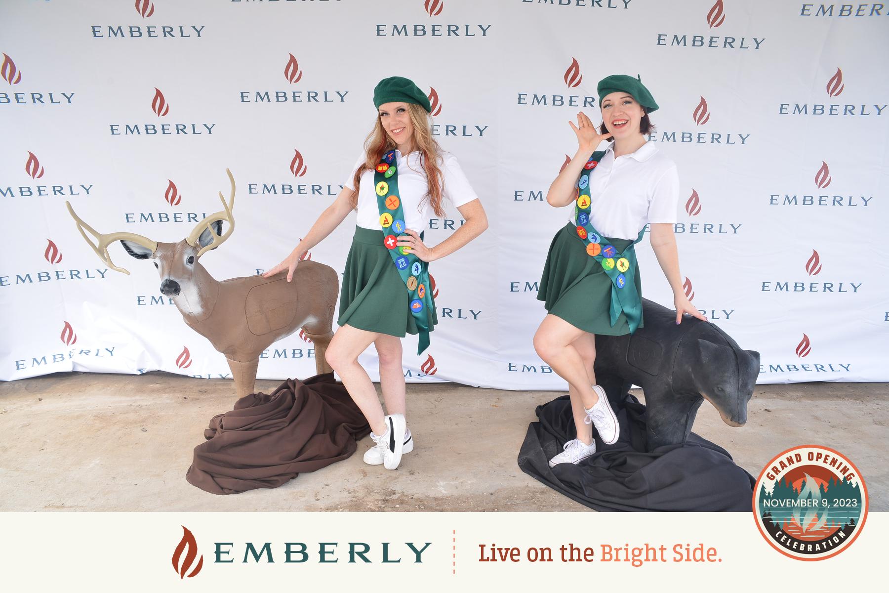 Two people in green outfits and sashes adorned with multiple badges stand on statues of a deer and bear, set against an "Emberly" backdrop with the text: "Emberly Live on the Bright Side," near Richmond, showcasing new homes.