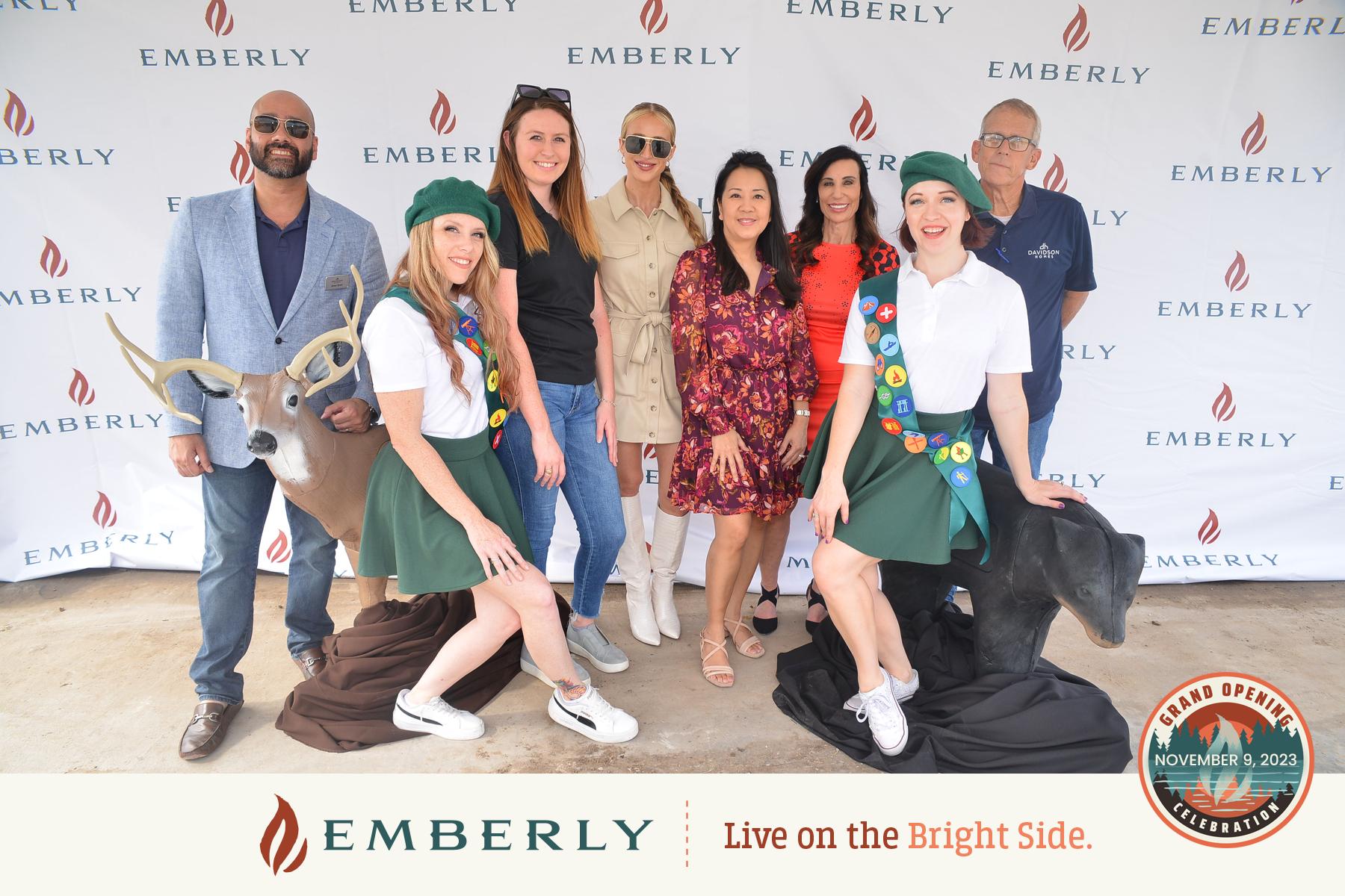 A group of people poses in front of a banner with "Emberly" written on it. Two women in scout uniforms stand by deer statues, celebrating the Grand Opening—November 9, 2023—of Emberly’s new homes near Rosenberg.