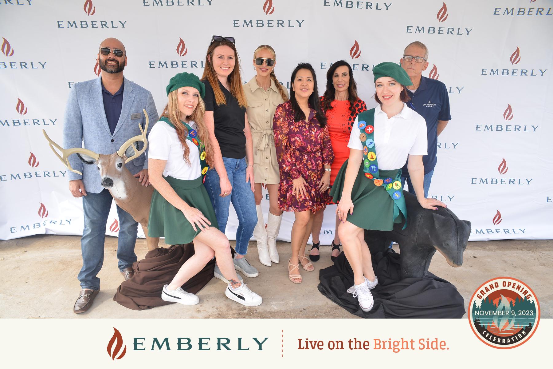 A group of people posing in front of an "Emberly" branded backdrop. Four men are standing alongside five women, with two of the women dressed in scout uniforms. The banner mentions a Grand Opening on November 9, 2023, near Rosenberg in Fort Bend County.