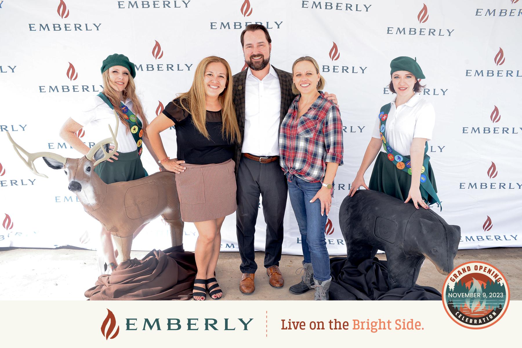 Five people stand smiling in front of an Emberly-themed backdrop, flanked by two deer sculptures. A logo featuring "Emberly" and "Live on the Bright Side" is at the bottom, along with a Grand Opening seal for this new homes master planned community near Rosenberg.