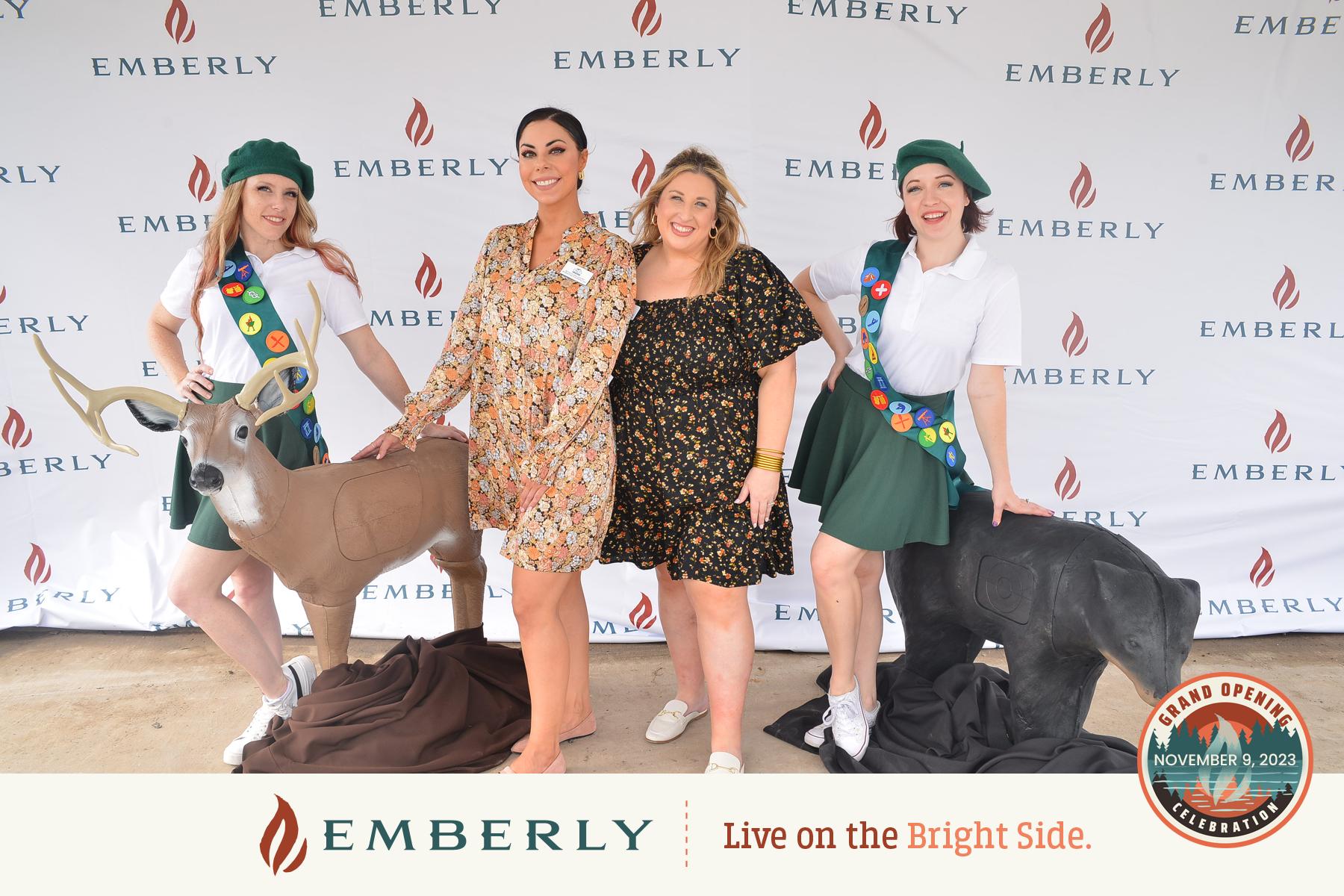 Four people pose in front of an Emberly banner featuring the slogan "Live on the Bright Side." Two individuals wear scout uniforms while the others wear floral dresses. In the foreground, two sculpted deer add charm, capturing a moment near Rosenberg in Fort Bend County