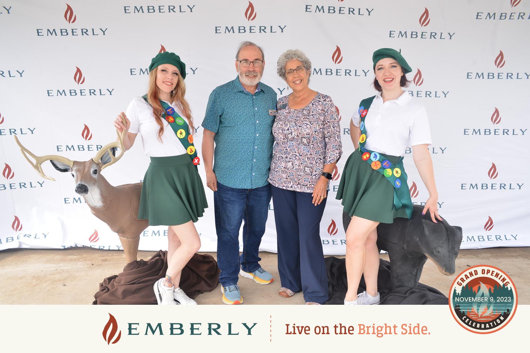 Four people pose in front of an Emberly backdrop, with two women dressed in themed costumes featuring sashes, green skirts, and berets. The backdrop and banner celebrate the grand opening of Emberly, a master planned community near Richmond.