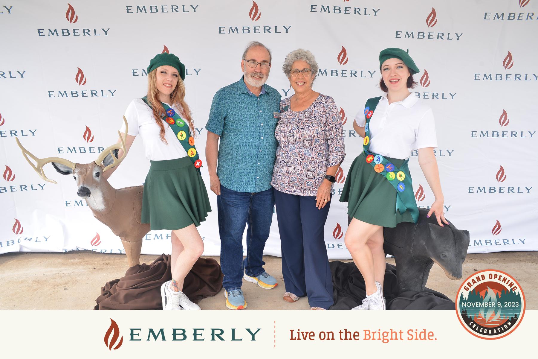 Two people stand between two costume mascots in scout uniforms in front of an Emberly-themed backdrop. The banner at the bottom reads "Emberly - Live on the Bright Side." and "Grand Opening November 5, 2023." Located near Richmond, Emberly is a vibrant master-planned community.