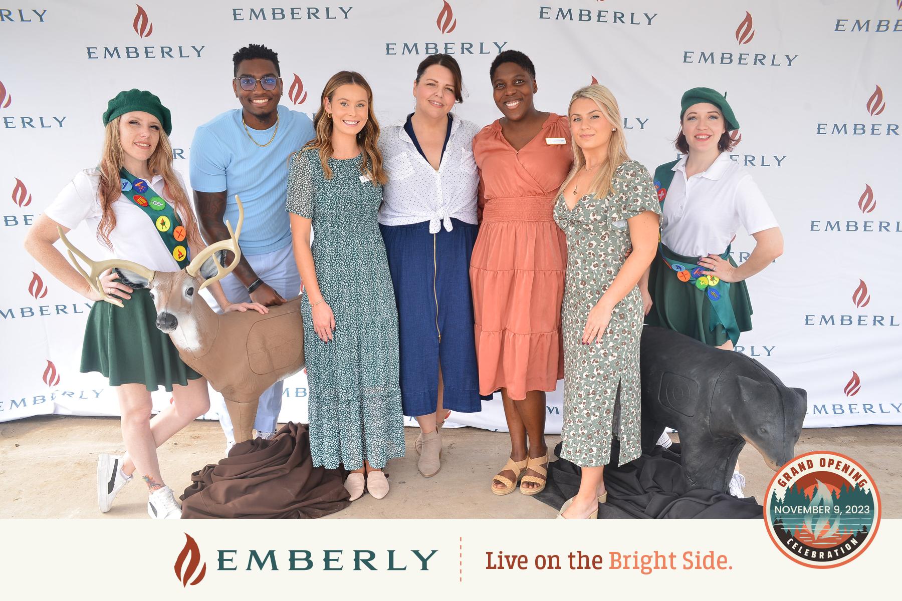 A group of seven smiling people stands in front of an Emberly backdrop with two deer statues. The banner reads, "EMBERLY, Live on the Bright Side," and highlights the grand opening date. Located near Richmond in Fort Bend County, these new homes promise a vibrant community experience.