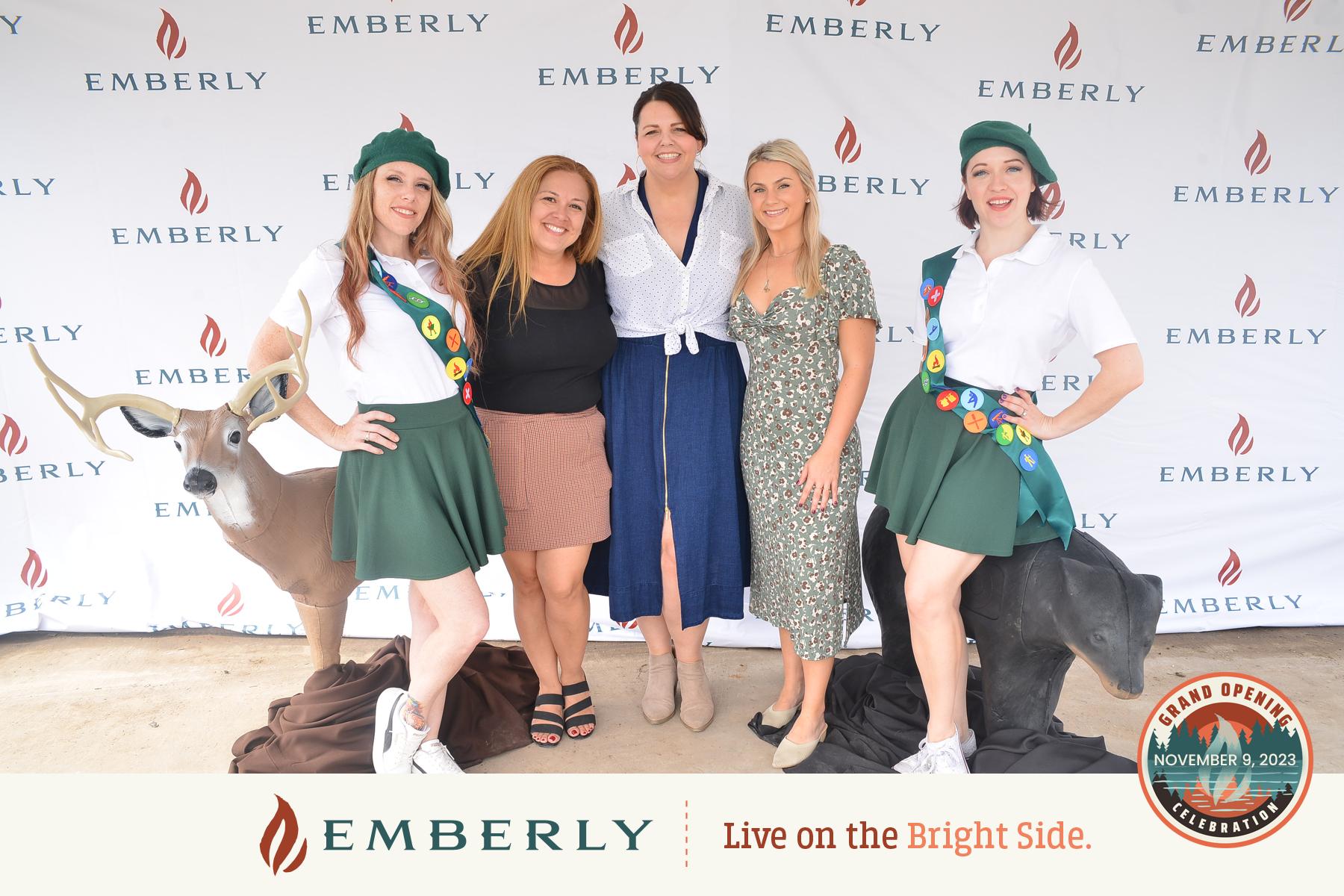 Five women smile and pose together in front of an Emberly step-and-repeat banner, with two deer statues beside them. The banner also features "Grand Opening November 9, 2023" text, celebrating the new master planned community near Rosenberg in Fort Bend County.