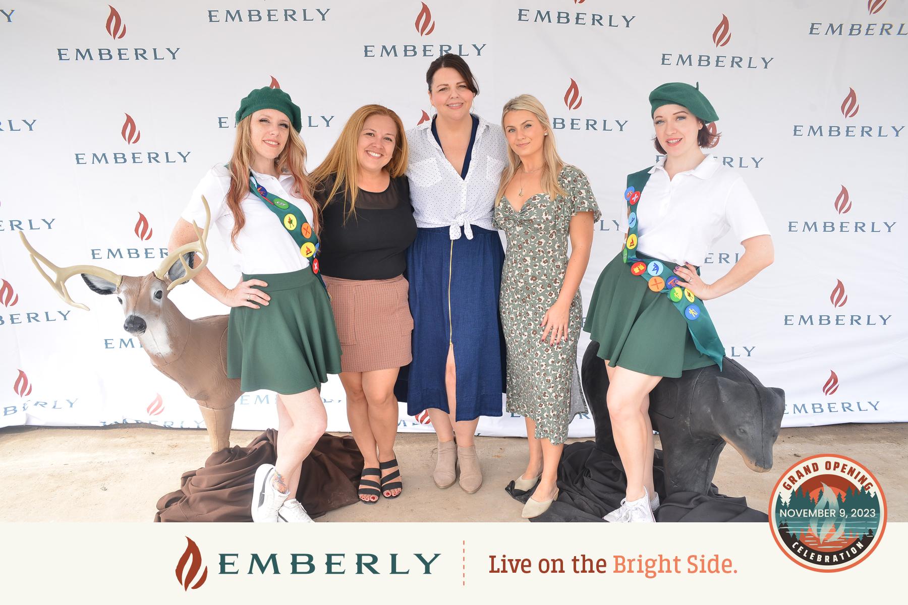 Five women pose for a photo in front of an "Emberly" backdrop, with two dressed as scouts. The text at the bottom reads "Emberly Live on the Bright Side" and shows details of a grand opening event near Richmond.