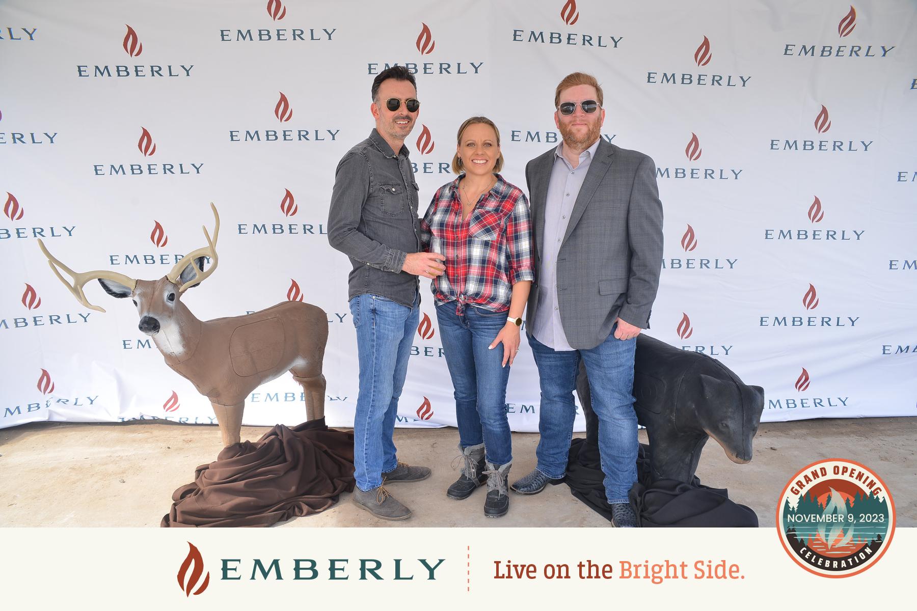 Three people stand in front of an "Emberly" branded backdrop, flanked by deer statues, at an event near Richmond with the sign "Grand Opening November 9, 2023.