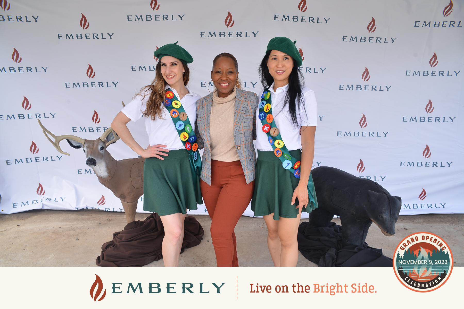 Three people stand smiling in front of an Emberly backdrop at an event near Rosenberg. Two individuals wear green uniforms with berets and sashes adorned with badges, while the third person in the center wears a plaid blazer, celebrating their involvement in a new master planned community.