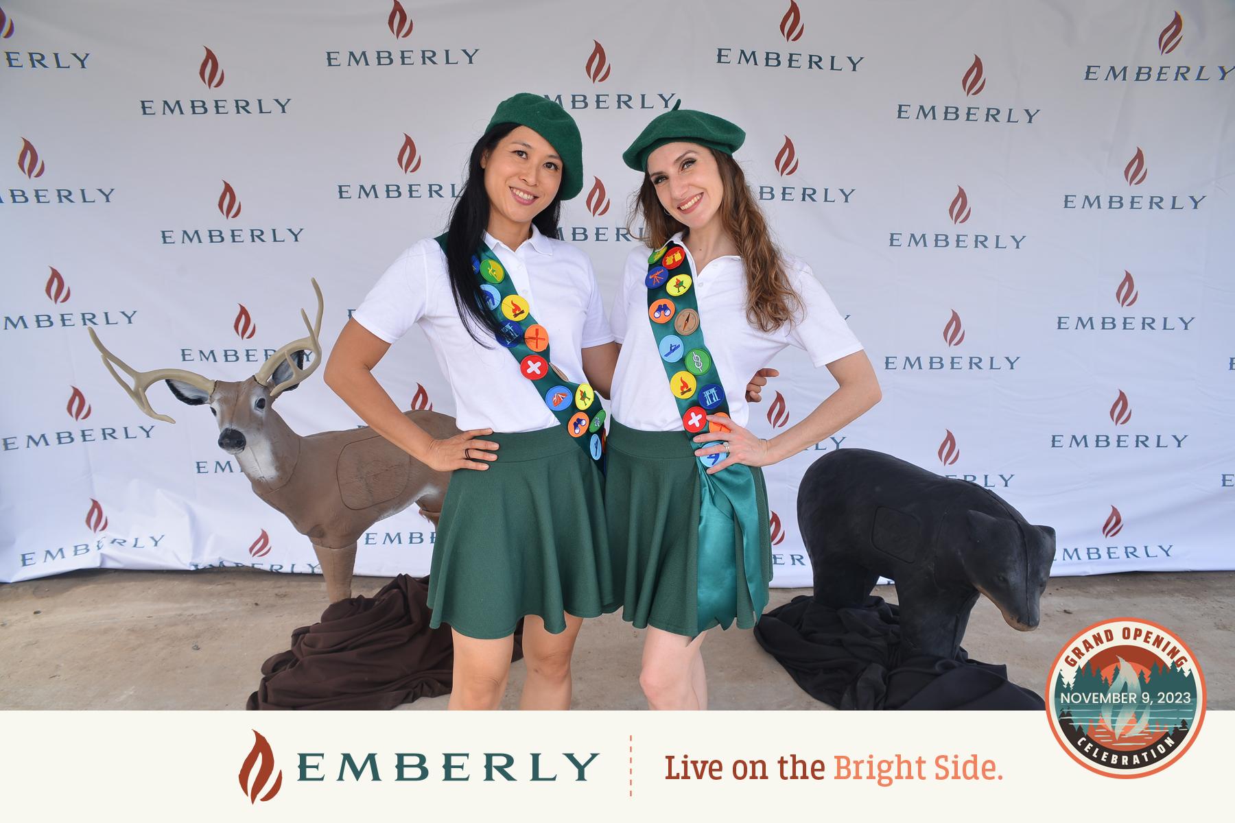 Two people dressed in scout uniforms pose in front of a backdrop with the Emberly logo near Richmond; deer statues are placed behind them. Text reads "EMBERLY Live on the Bright Side. Grand Opening" with the date November 3, 2023.