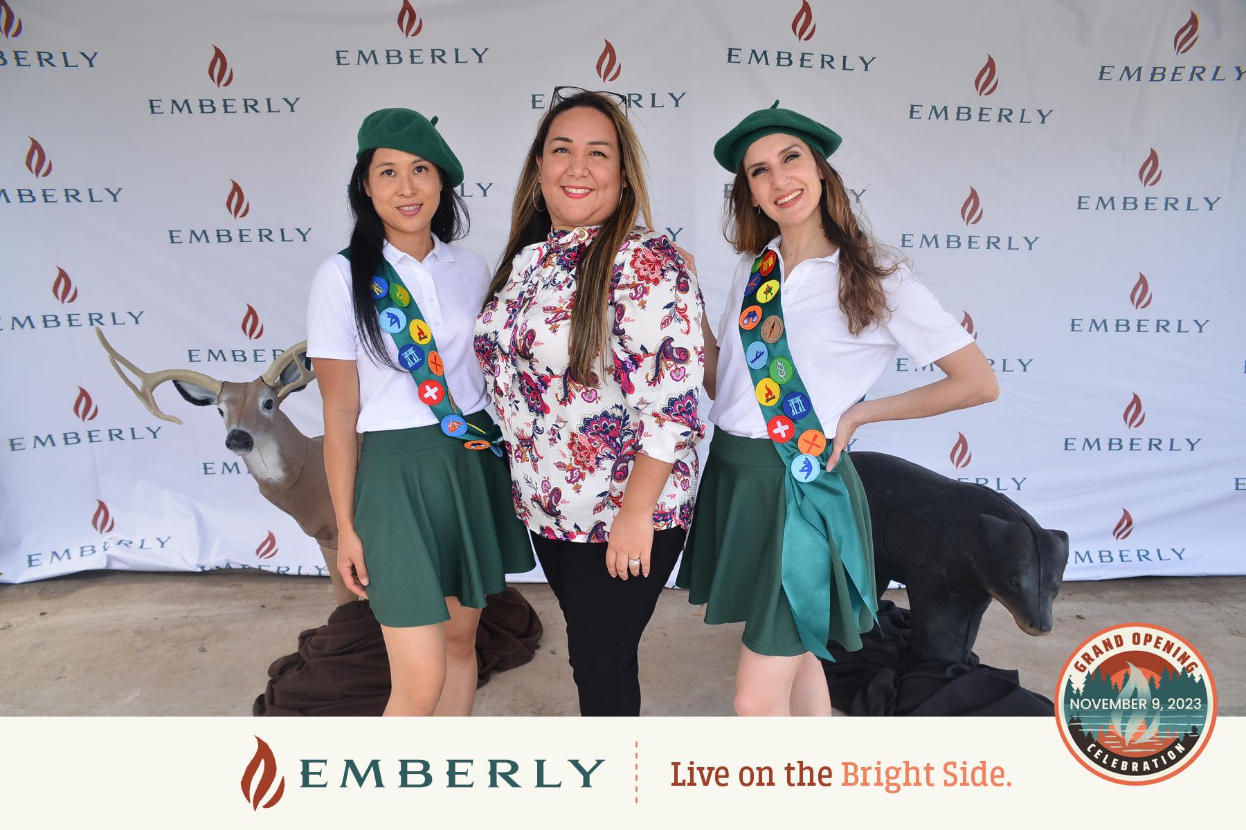 Three women pose for a photo at an Emberly event promoting new homes near Rosenberg. Two women wear green berets and skirts with sashes, while one wears a floral blouse. The backdrop features the Emberly logo and event details, highlighting its connection to Fort Bend County living.