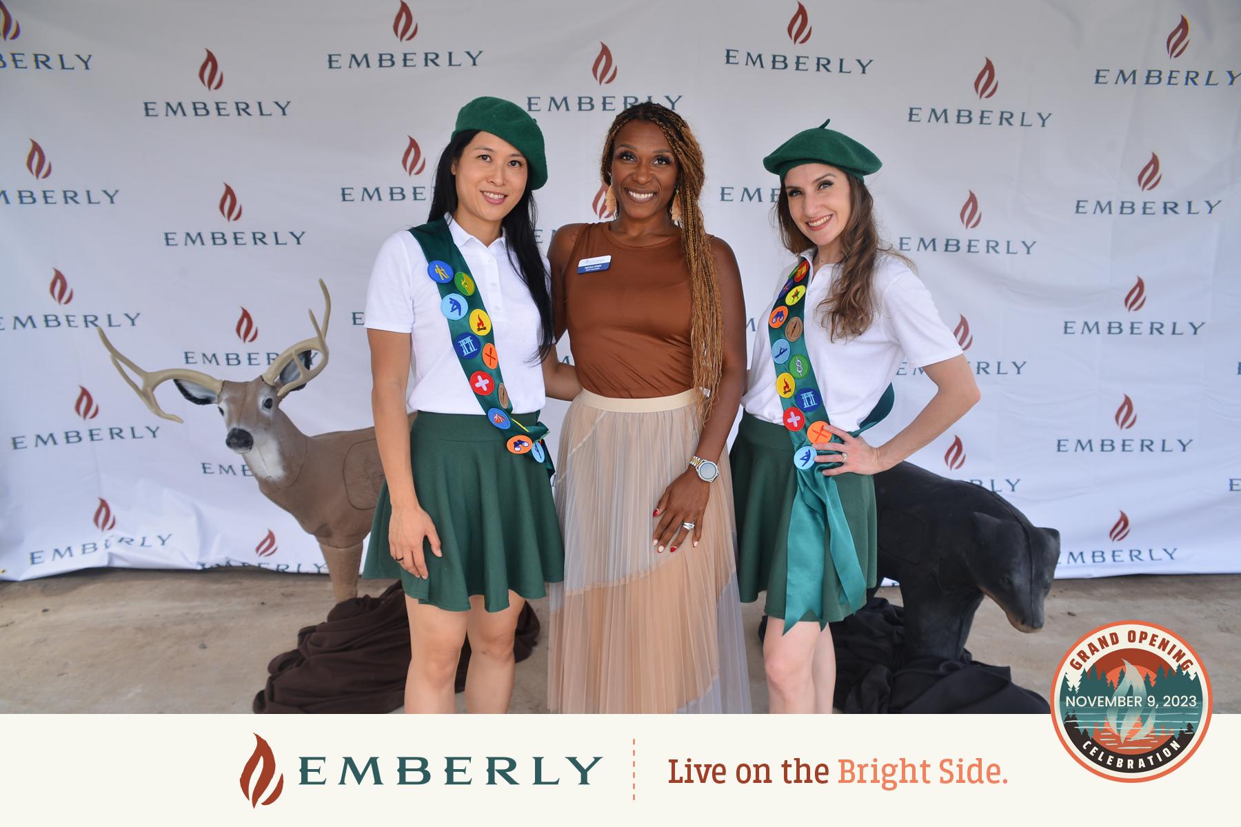 Three women pose in front of an Emberly backdrop at an event. Two are dressed in scout uniforms with green berets and decorated sashes, while the third is in a brown top and tan skirt. The scene highlights the community spirit at Emberly, a master planned community showcasing new homes.