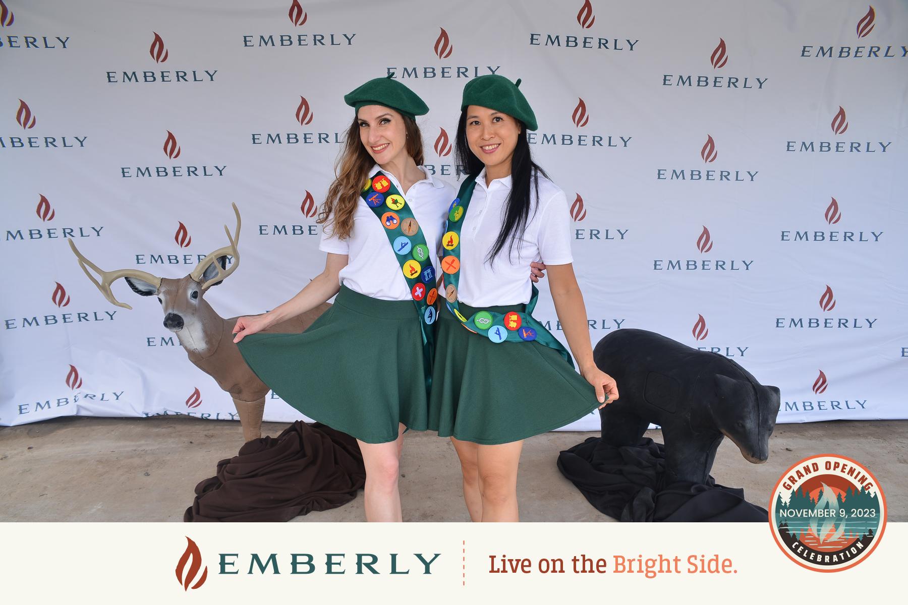 Two women dressed in green skirts and hats pose in front of an Emberly backdrop near Richmond. A deer and a bear statue can be seen on either side of them. A banner reads "Emberly Live on the Bright Side.