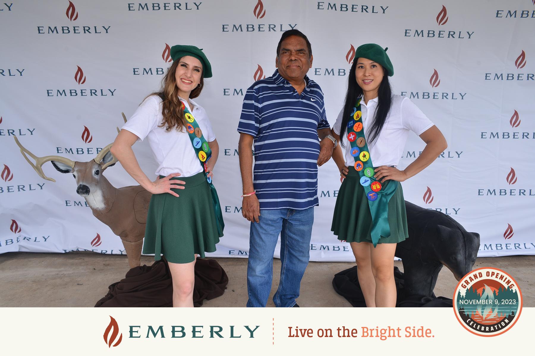 A man stands between two women dressed as scouts in front of an Emberly backdrop, showcasing a master planned community near Richmond. A deer model is visible in the background. The Emberly logo and "Live on the Bright Side" slogan are at the bottom, promoting Fort Bend County