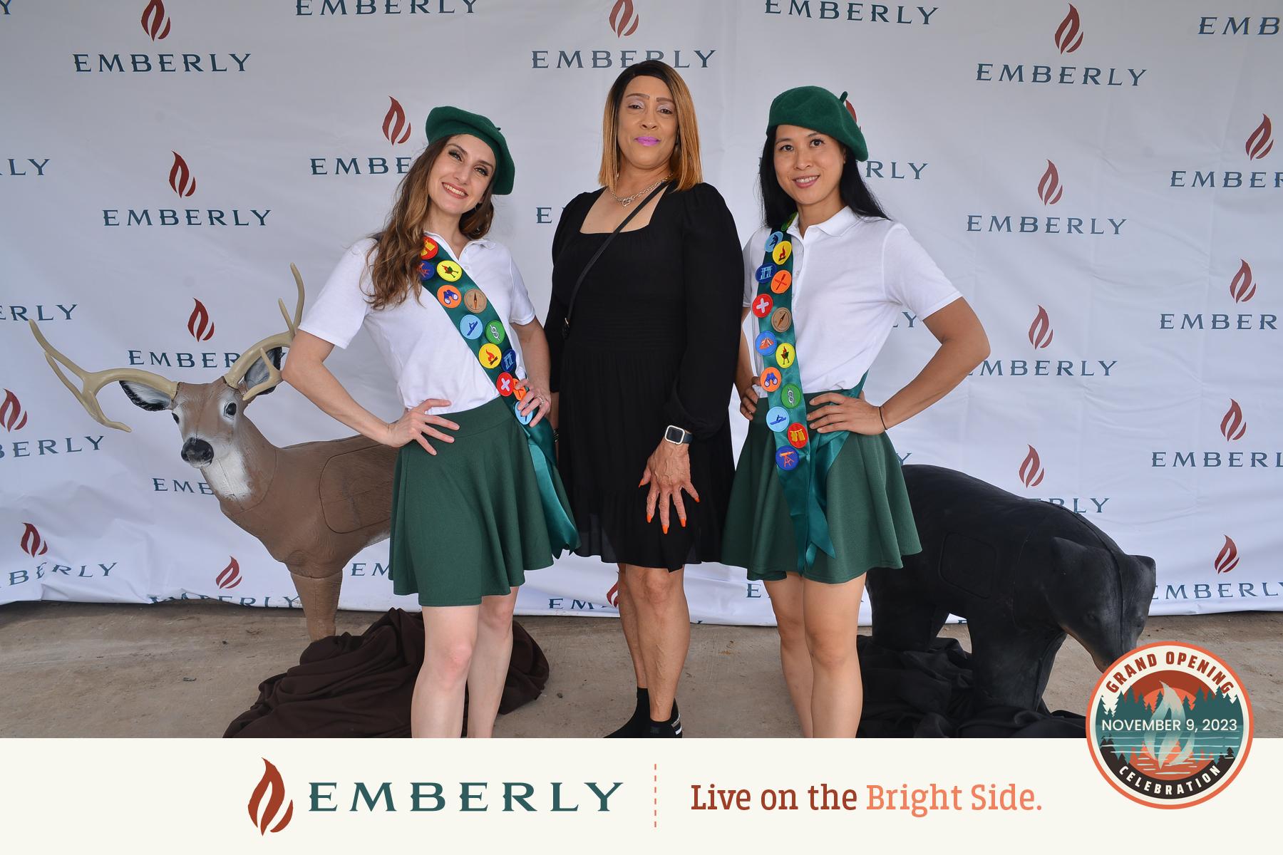 Three women pose in front of an Emberly-branded backdrop at an event on November 5, 2023. Two women wear matching uniforms with green skirts and berets, while the woman in the center wears a black dress. The event, held near Richmond and promoting new homes, was a resounding success.