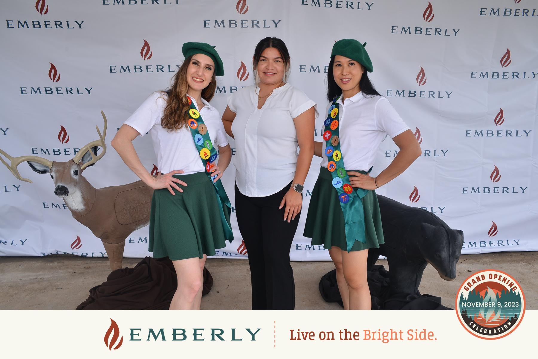 Three women pose in front of an Emberly backdrop, capturing the essence of this master planned community. Two wear green berets and scout-like uniforms, while the third is in a white top and black pants. A fake deer and a seal add whimsical charm to the background.