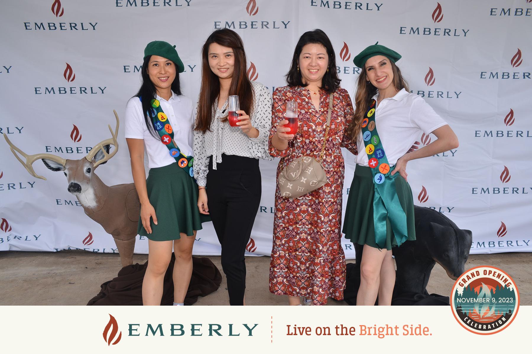 Four women smiling and holding drinks stand in front of an "Emberly" branded backdrop. Two are dressed in scout uniforms, with a staged deer visible behind them. Text reads, "Live on the Bright Side." Enjoy life near Richmond and Fort Bend County with Emberly