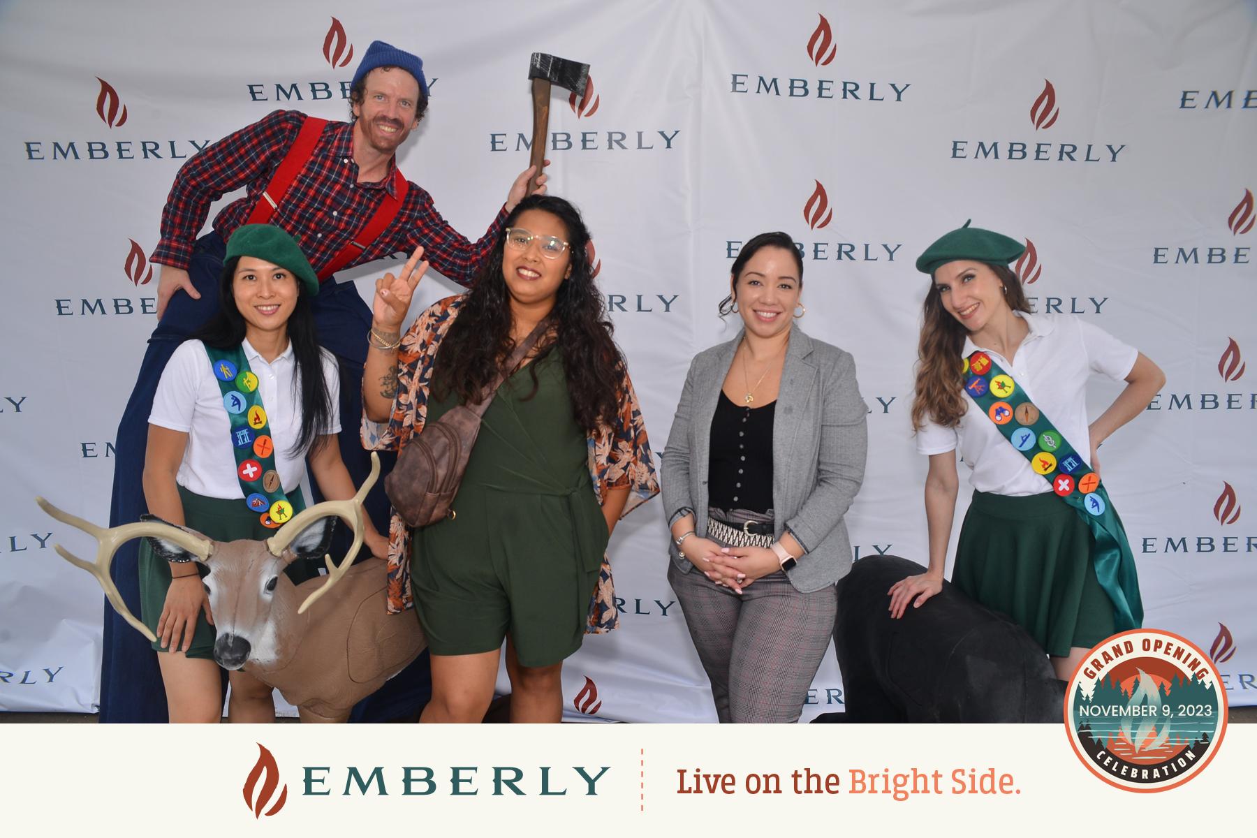 Five people posing in front of a backdrop with "Emberly" logos. Two women are dressed as Girl Scouts, one person is dressed as a lumberjack, and two others are in business attire. A stuffed deer is in the foreground, symbolizing the rustic charm of Emberly’s new homes within this master planned community.