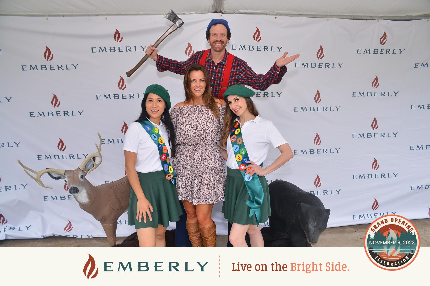 Four people posing in front of an "Emberly" branded backdrop at an event in Fort Bend County. One person is holding an axe, two are dressed in green, and one in brown boots is in the center, smiling. A stuffed deer is on the side, highlighting the charm of this master planned community.