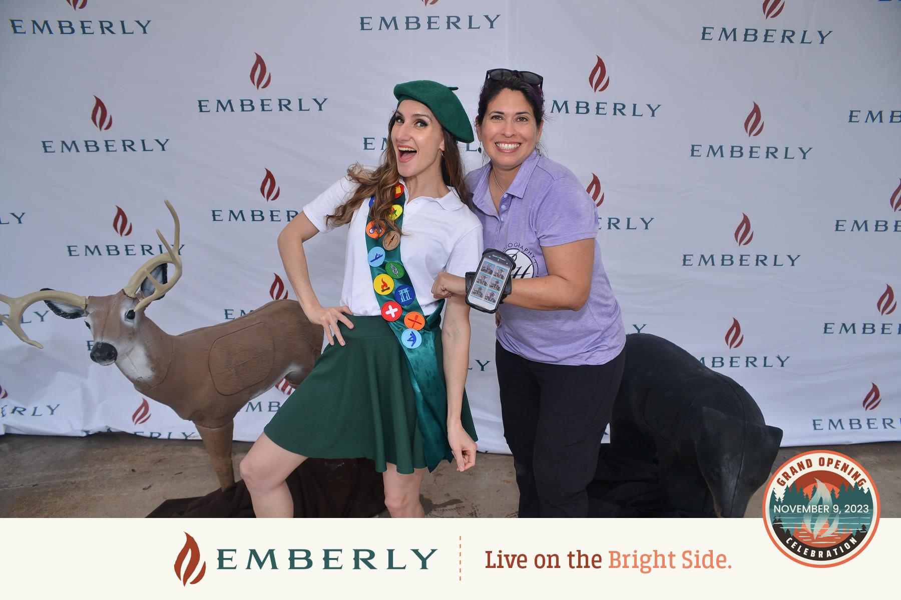 Two people pose in front of an Emberly backdrop, one wearing a green beret and sash with badges, the other holding a mobile phone. A deer and a fur-covered bike are also in the scene at this whimsical event near the master planned community known for its new homes near Rosenberg.