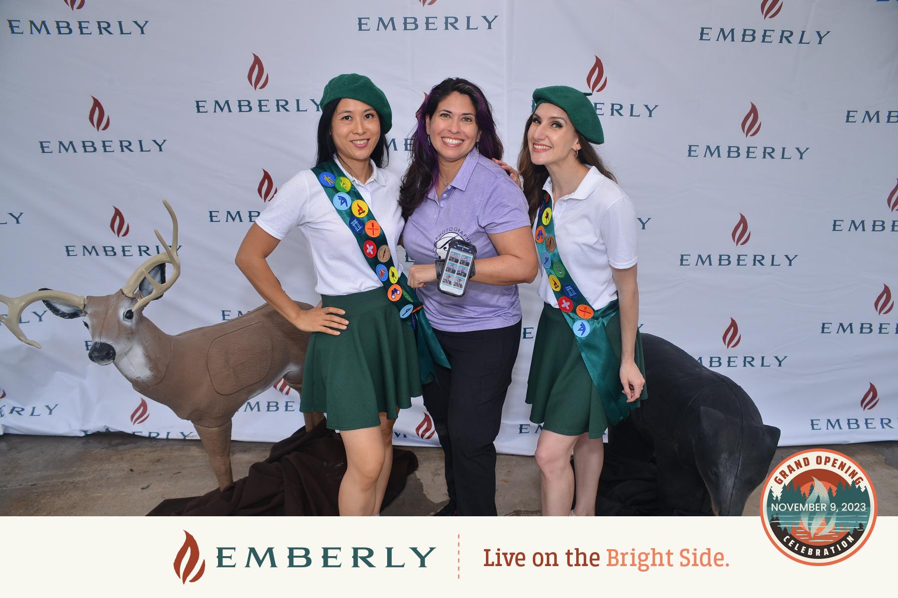 Two people in scout uniforms pose with a woman holding a device in front of an Emberly branded backdrop with a deer mannequin. Text on the banner reads, "Emberly Live on the Bright Side," showcasing new homes in a master planned community near Richmond.