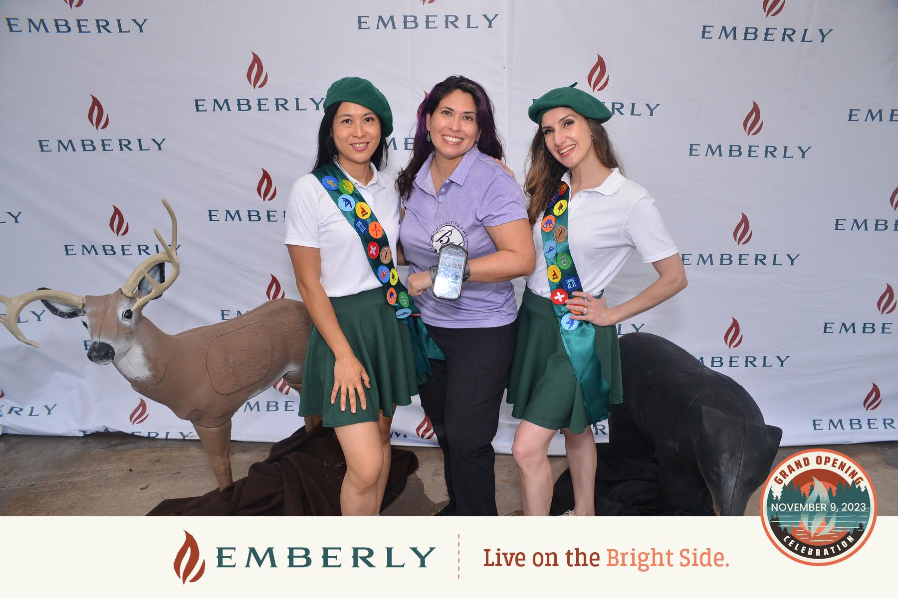  Three women posing together in front of an Emberly backdrop, two wearing green berets and sashes, one holding a spray bottle. A deer statue is beside them. Text reads "Grand Opening" and "November 9, 2023". Located near Rosenberg in a master planned community.
