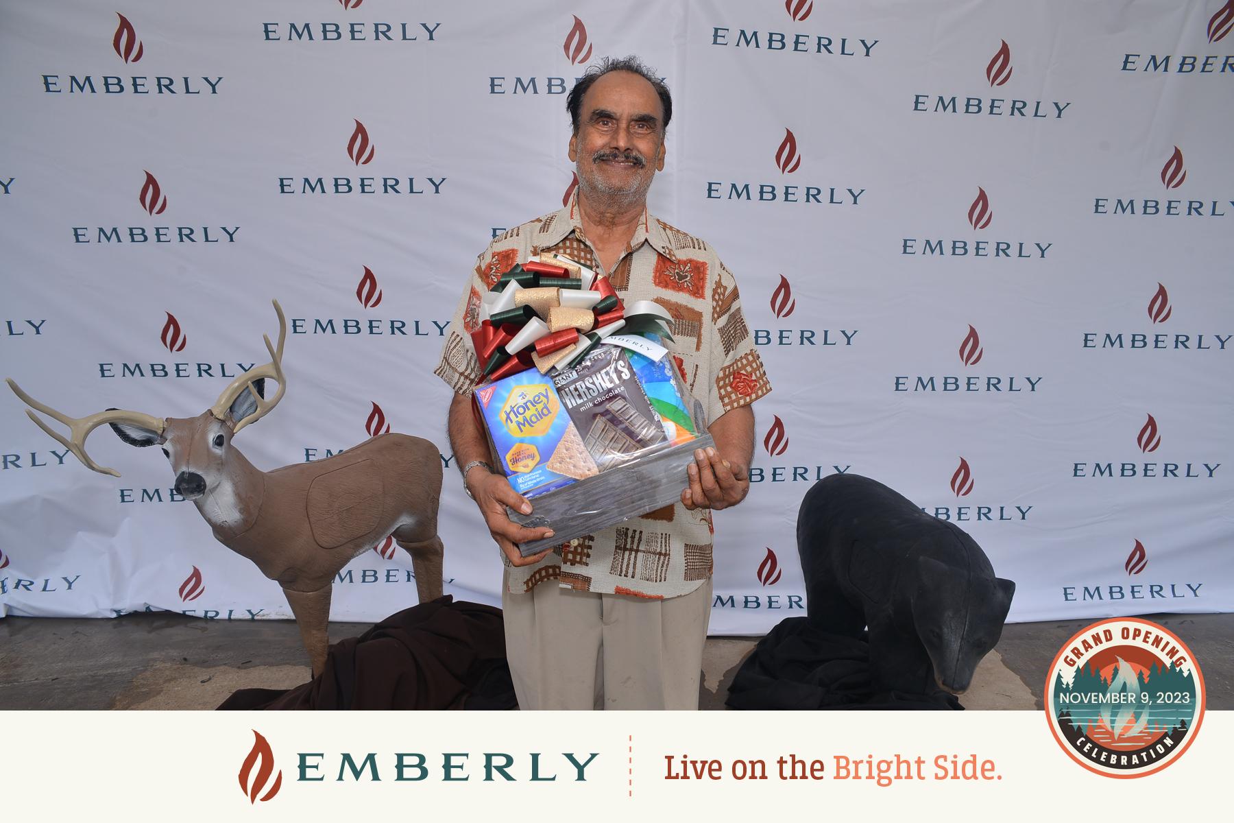 A man stands in front of an Emberly branded backdrop, holding a stack of household items. Decorative figures of a deer and a bear are visible beside him. The backdrop mentions the grand opening on November 9, 2023, in Fort Bend County