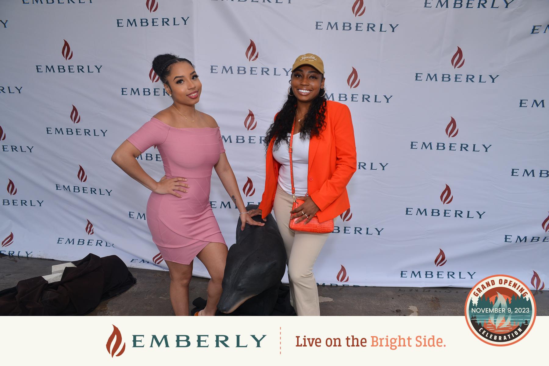 Two women pose in front of an Emberly branded backdrop, epitomizing the charm of this master-planned community. The woman on the left stuns in a pink dress, while the woman on the right captivates in an orange blazer and white pants. Event signage is visible at the bottom.