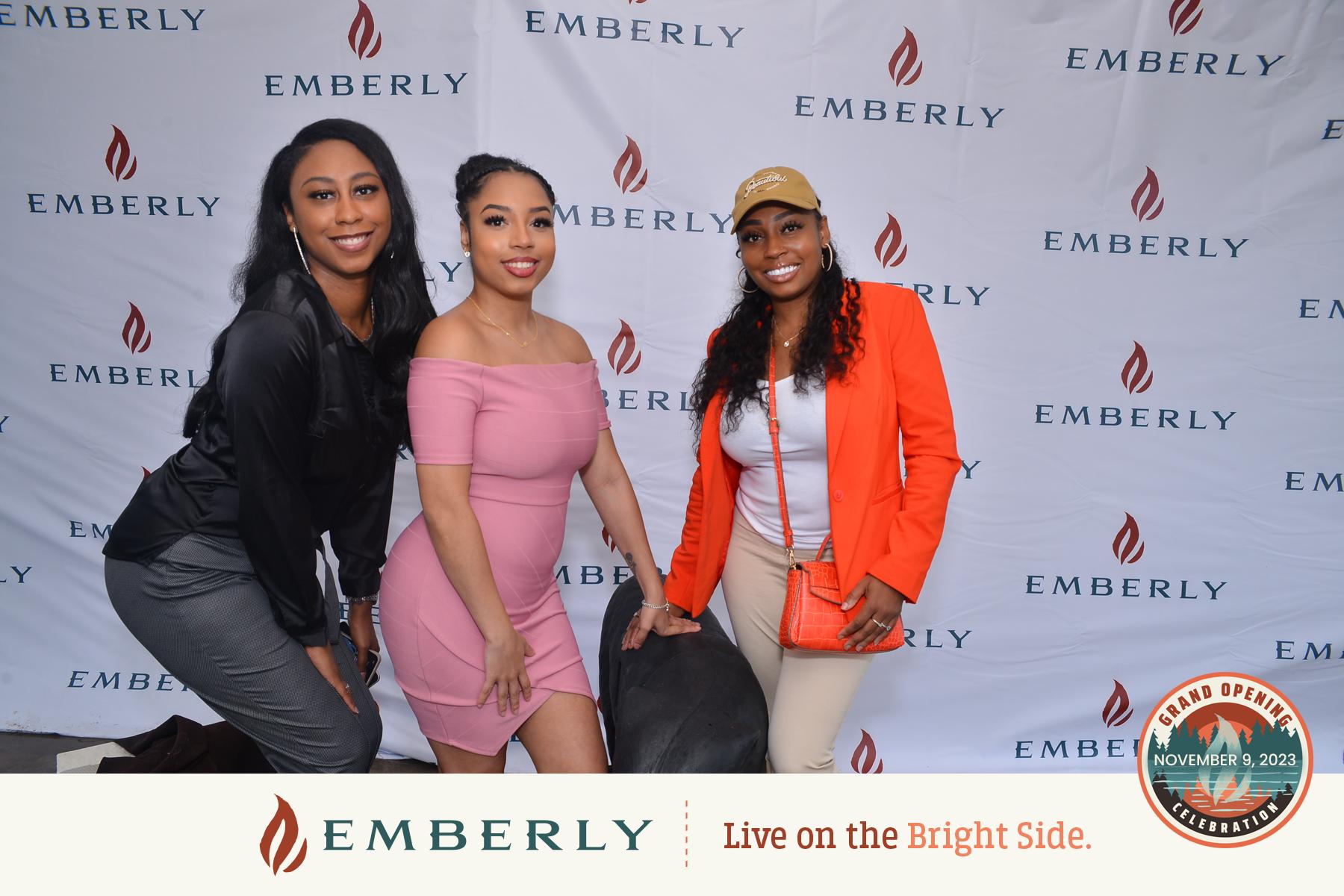 Three women smiling and posing in front of a backdrop with "EMBERLY" logos. One is standing, and two are seated. A circular sign with "Grand Opening Celebration, November 9, 2023" is in the bottom right corner, marking the debut of Emberly