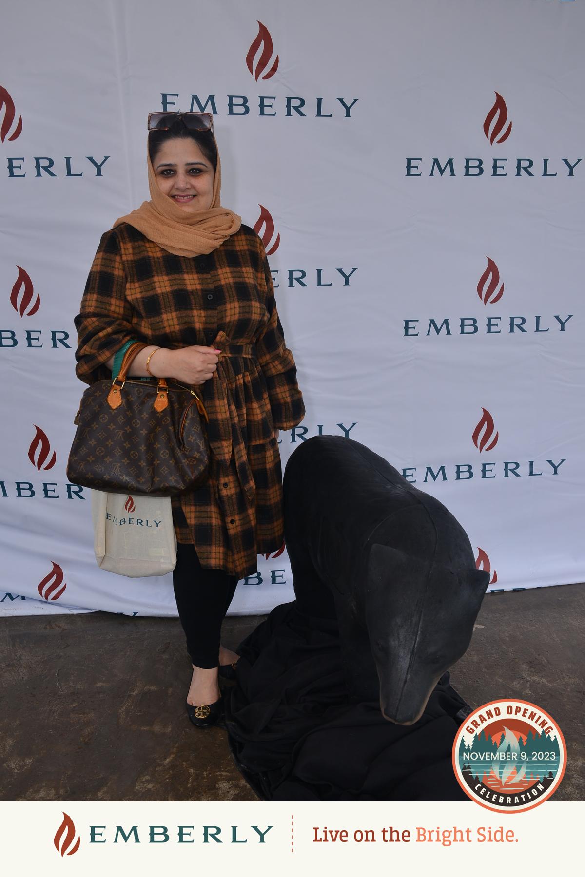 A woman in a checkered dress stands next to a black statue, holding a handbag and a tote bag, on a backdrop featuring the "EMBERLY" logo. Text on the backdrop mentions the grand opening of Emberly, a master planned community near Rosenberg.