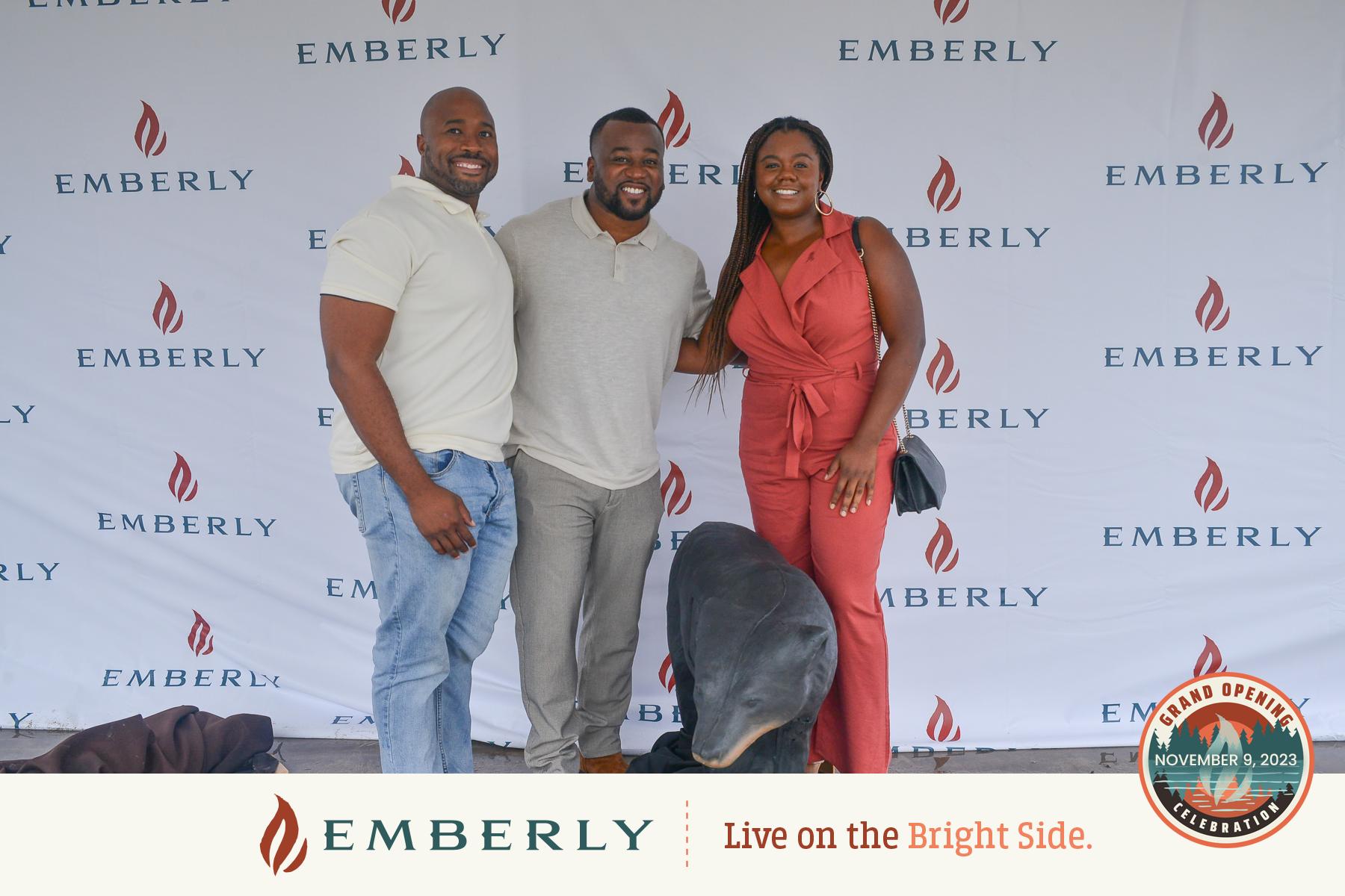 Three people are standing and smiling in front of an Emberly-branded backdrop, near Rosenberg. One person is petting a dog. The backdrop includes a logo and text promoting the grand opening of this master planned community on November 9, 2023.