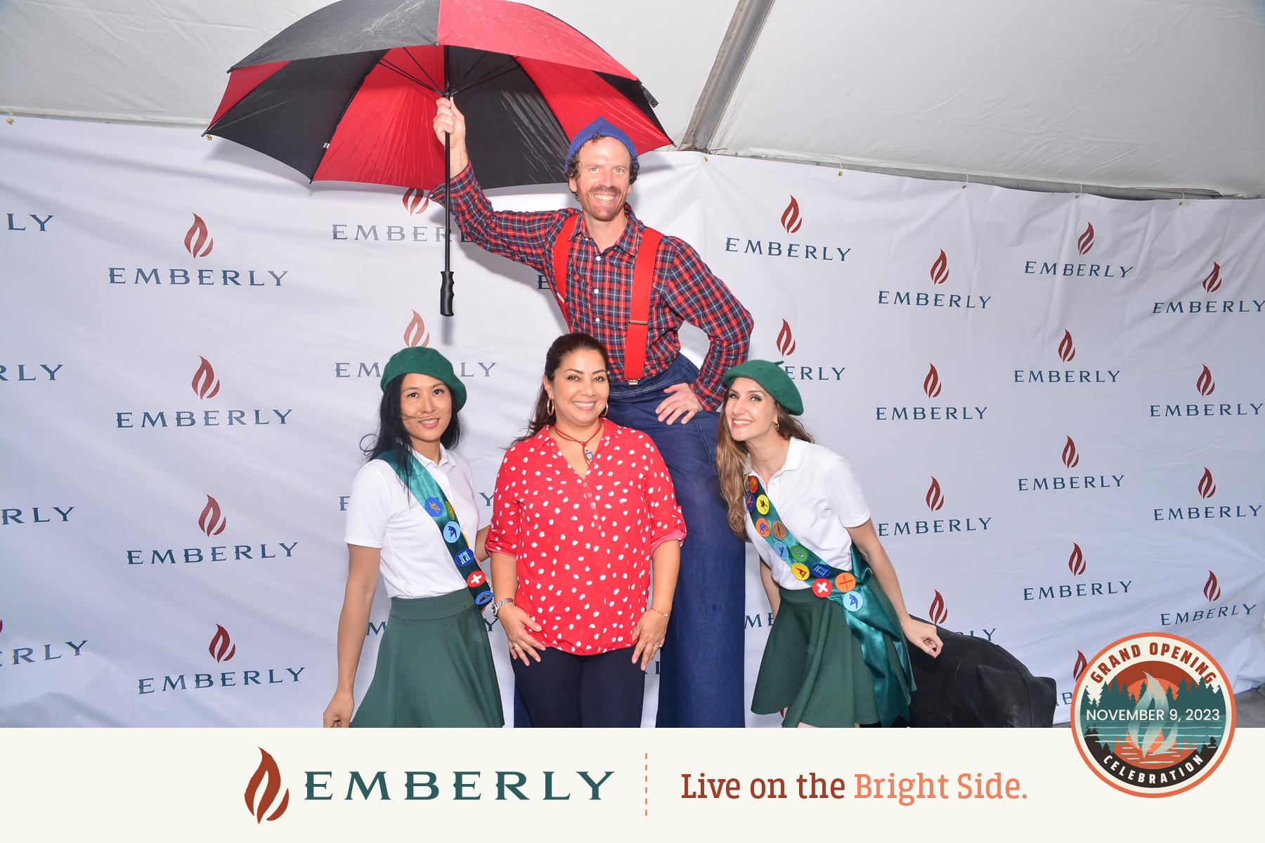 Four people posing together in front of a branded backdrop near Rosenberg. A man in a red plaid shirt holds an umbrella, standing behind three women—two in green scout uniforms and one in a red polka dot blouse, celebrating the opening of new homes in a master-planned community.