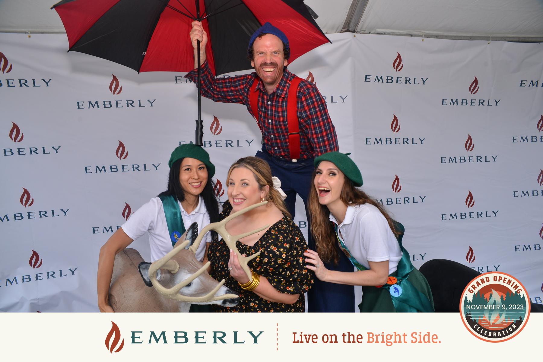 Four people posing for a photo with a reindeer antler prop. One person stands behind holding a red and black umbrella. The backdrop displays the Emberly logo and text reading "Grand Opening November 9, 2023," near Richmond.