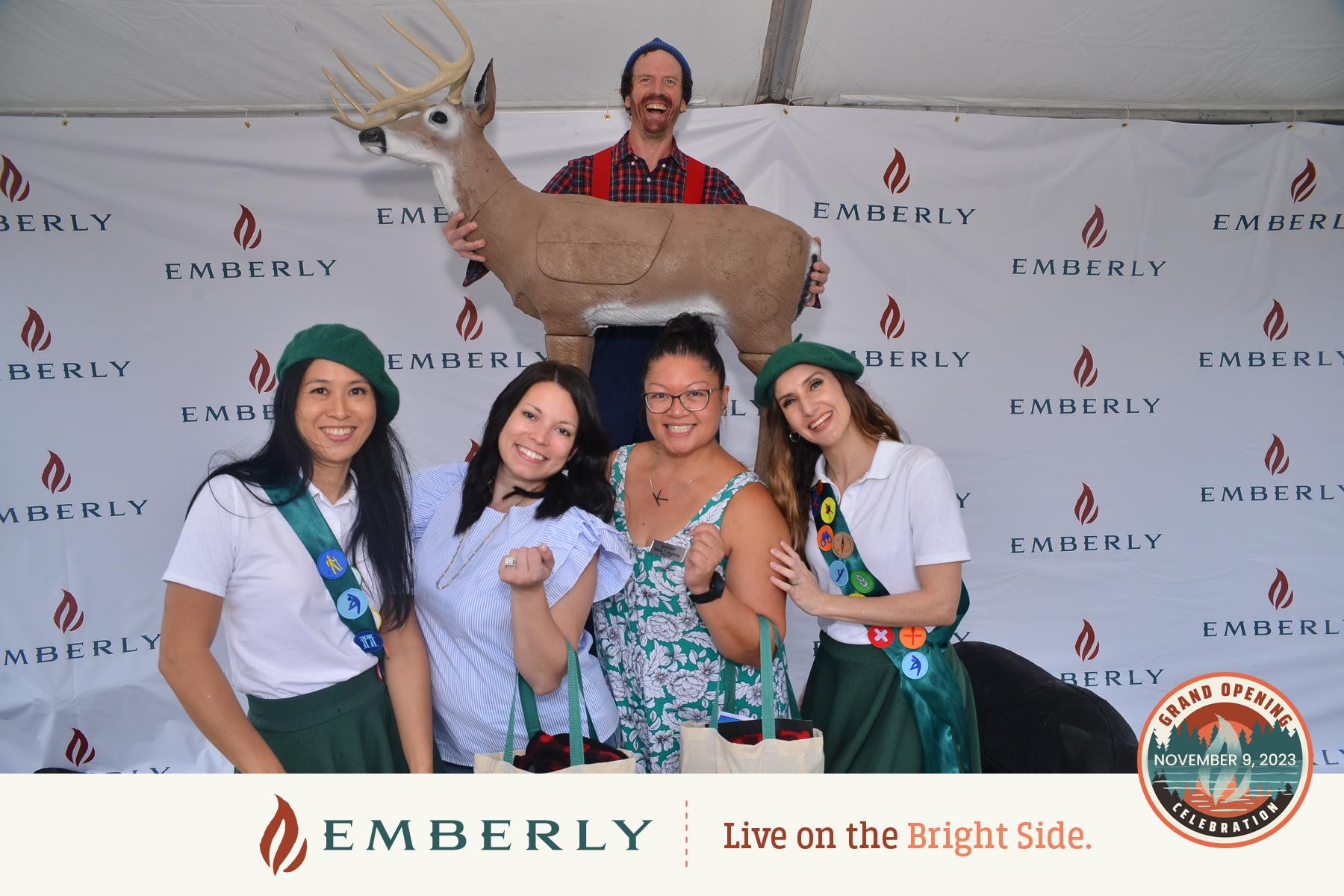 A group of four people, three dressed in green scout uniforms, pose in front of an Emberly backdrop while a man stands behind them holding a deer statue. A sign reads "Grand Opening Celebration November 9, 2023" for the master planned community with new homes near Rosenberg.