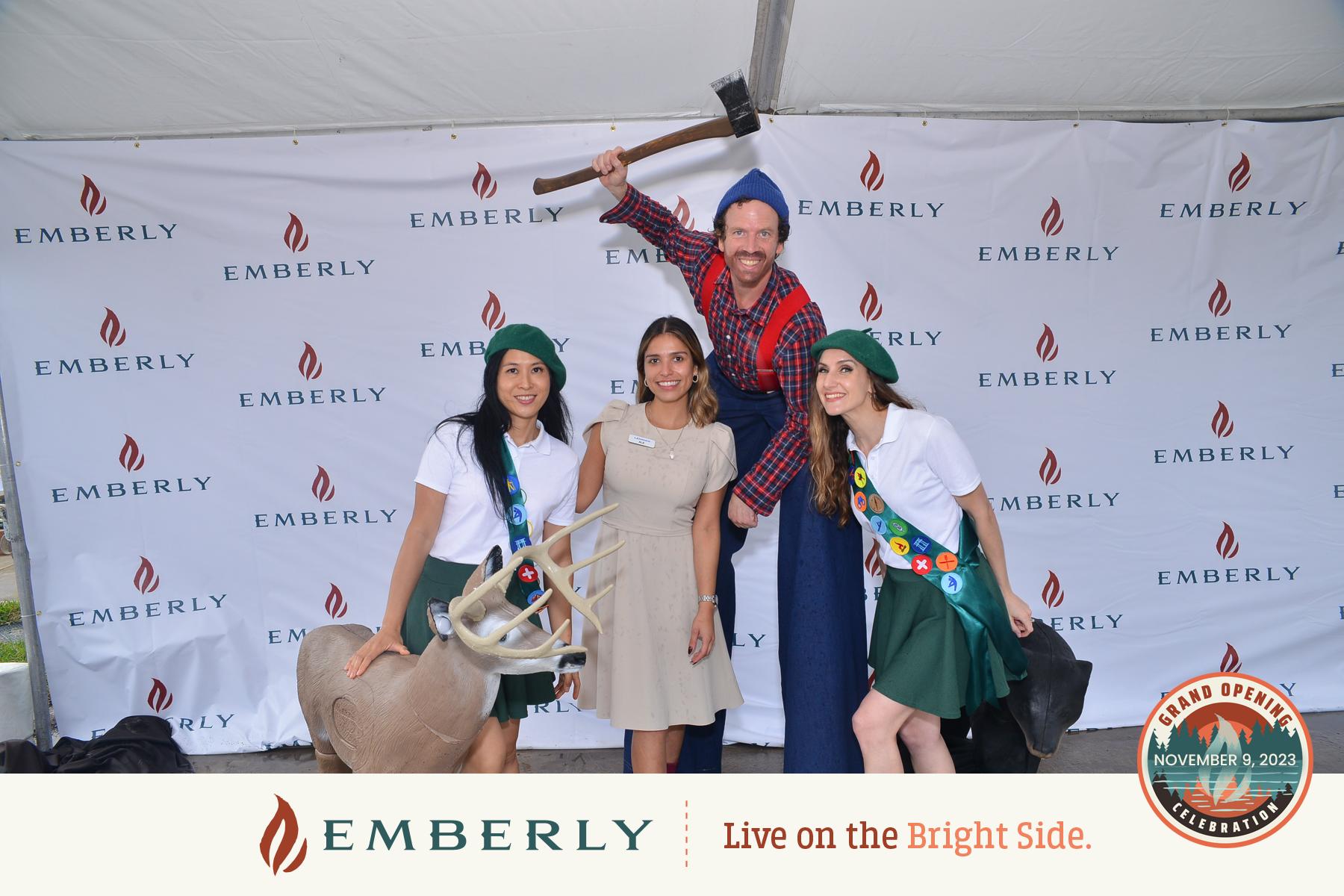 Four people posing in front of an Emberly-branded backdrop near Rosenberg. One person is holding an axe, while another stands beside a deer statue. This event marks the grand opening of new homes on November 3, 2023.